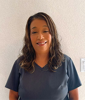 Smiling woman wearing dark gray scrubs against a light textured wall.